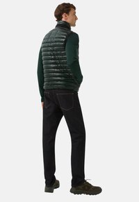 Mann steht mit den Händen in den Taschen einer dunkelgrünen gesteppte Weste, trägt dunkelblaue Jeans, ein grünes langärmliges Hemd und grüne Wanderschuhe auf weißem Hintergrund.