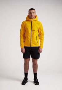 Gul vandtæt jakke med hætte, frontlynlås, sorte detaljer og et logo. Kombineret med sorte shorts og sorte atletiske sko.