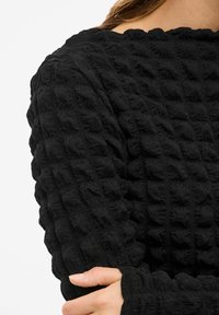 Maglione nero con texture e motivo trapuntato, dotato di scollatura tonda e maniche lunghe. Il tessuto appare morbido e leggermente patternizzato.