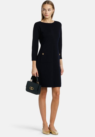 Marineblaues, figurbetontes Kleid mit langen Ärmeln, ausgestattet mit zwei Taschen mit goldenen Knöpfen, kombiniert mit einer schwarzen Handtasche und goldenen flachen Schuhen.