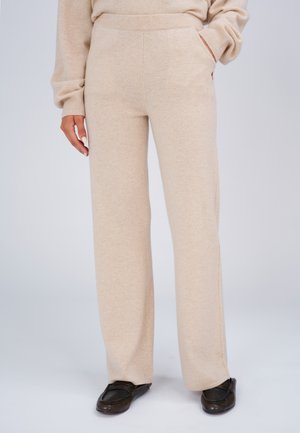 Beige weite Strickhosen aus weichem Material, getragen mit einem passenden Pullover und schwarzen Slipper-Loafern, eine Hand in der Tasche.