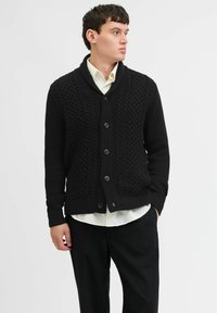 Schwarze Strickjacke mit strukturiertem Muster, Schalkragen und sechs Knöpfen. Getragen über einem weißen Hemd und kombiniert mit schwarzen Hosen.