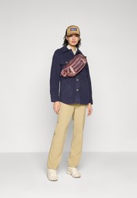 Marine fleecejakke, beige bukser og kremfargede joggesko. Lilla mageveske med "Patagonia"-logo; justerbar stropp og slankt design.