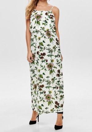Femme portant une robe longue blanche avec des imprimés floraux verts et marron et des chaussures à talons hauts noires, debout devant un fond uni.