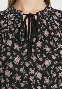 Blouse noire à motifs floraux roses, avec col à volants et fermeture à nouer à l'avant. Fabriquée en tissu léger et texturé.