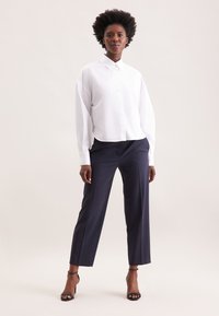 Donna in piedi che indossa una camicia bianca a maniche lunghe con bottoni, pantaloni cropped blu scuro e tacchi a caviglia neri su uno sfondo neutro.
