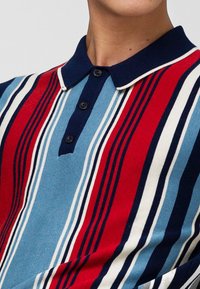 Randig polo-shirt i röd, blå och vit med en marinblå krage, med en knappslå med tre svarta knappar och textilknittrande tyg.