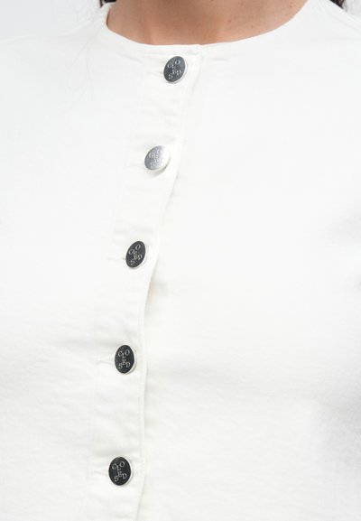 Λευκό ύφασμα τοπ με στρογγυλή λαιμόκοψη, κάθετη κλείσιμο με κουμπιά και έξι μεταλλικά κουμπιά με λογότυπο σε κάθε ένα.