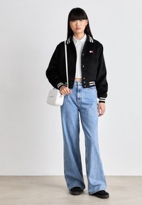 Sort varsity-jakke med hvid stribet krave, hvid skjorte, lyseblå brede jeans og en lille hvid crossover-taske.