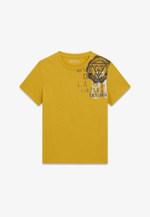 T-shirt jaune moutarde à manches courtes avec col rond et texte imprimé noir ainsi qu'un emblème sur le haut côté gauche de la poitrine indiquant "Guess Los Angeles California."