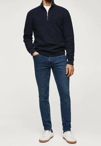 Pull en maille bleu marine avec demi-zip, associé à un jean bleu ajusté et des baskets blanches à semelles beige, se tenant sur un fond neutre.