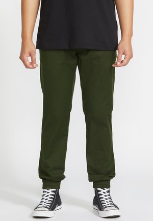 Homme portant un pantalon jogger vert olive, des baskets montantes noires avec des lacets blancs et un t-shirt noir, debout devant un fond uni.