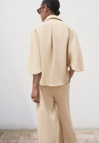 Beige, leichtes Oberteil mit einem umklappbaren Kragen und weiten Ärmeln, kombiniert mit passenden weiten Hosen. Glatte, leichte Stoffstruktur.