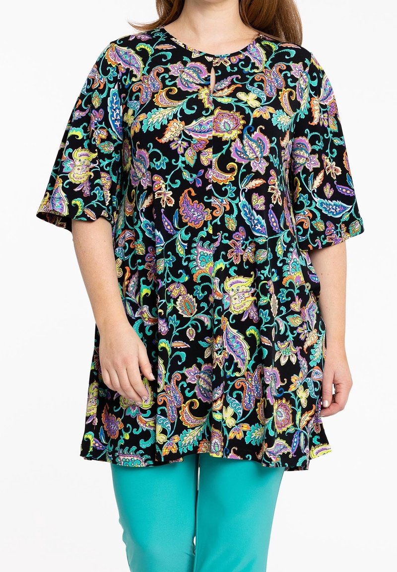 Femme portant une tunique noire ample avec des motifs cachemire colorés et des leggings bleu sarcelle, debout les mains détendues le long du corps.