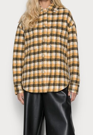 Mujer vistiendo una camisa de cuadros amarilla, marrón y blanca con botones, combinada con pantalones de cuero negros de pierna ancha.