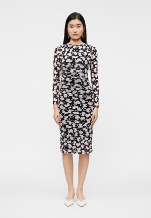 Μαύρο φόρεμα με μακριά μανίκια και ροζ και λευκό λουλουδάτο μοτίβο, εφαρμοστό σιλουέτα, μήκος μέχρι τη μέση της γάμπας, σε συνδυασμό με λευκά παπούτσια μυτερά.