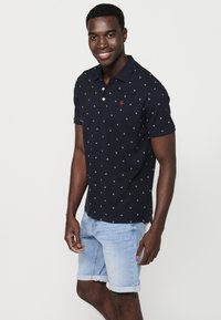 Námornícky polo tričko s malými bielymi a červenými geometrickými vzormi, so zapínaním na dva gombíky pri krku. Kombinované so svetlomodrými džínsovými šortkami, s olemovaným okrajom.