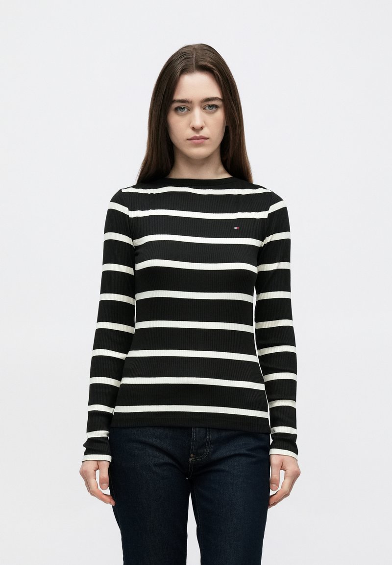 Must triiksärk valgete horisontaalsete triipudega, sooniline tekstuur, ümarsääriline kaelus ja väike logomärk rinnal, sobitatud tumedate teksadega.