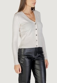 Cardigan a V, con maniche lunghe, in maglia beige chiaro con orlo a costine e cinque bottoni metallici sulla parte frontale; abbinato a pantaloni in pelle nera a vita alta.