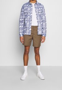 Man som bär en blå mönstrad jacka över en vit t-shirt, bruna shorts, vita strumpor och vita sneakers, stående mot en plain ljusgrå bakgrund.