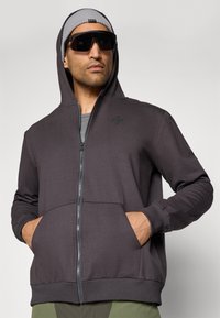 Sivá zip-up kapuca s prednou kapsou a rebrovanými manžetami. Nosí sa so sivým čiapkom a tmavými slnečnými okuliarmi. Hladká textúra látky.