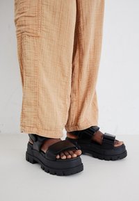 Black chunky sandals with a thick sole, wide strap design, and adjustable buckle, paired with loose light beige textured pants.