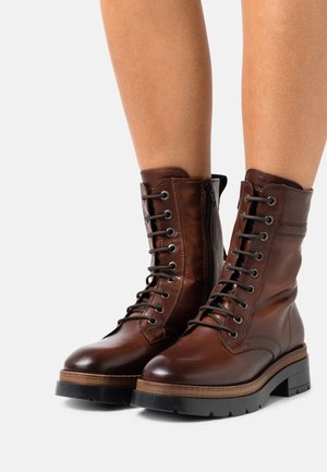 Brune læder ankelstøvler med snørebånd og chunky sorte såler, båret på bare ben mod en hvid baggrund.