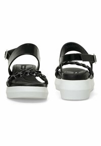 Sandalias de plataforma negras con tiras trenzadas, hebilla en el tobillo y suela blanca. Textura suave con un diseño moderno.