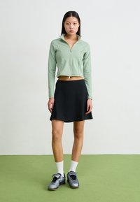 Zaļa īsā jaka ar rāvējslēdzēju, melna plisētā svārku, pelēki sporta apavi un balti zeķes, uz zaļa grīdas fona.