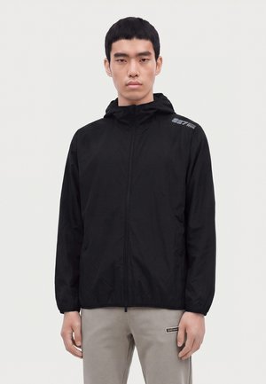 Homme portant une veste à capuche noire zippée et un pantalon beige, debout devant un fond clair uni.