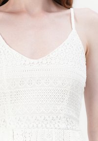 Weißes Kleid mit Spitzenmuster, V-Ausschnitt und dünnen Trägern, mit strukturierten Bereichen, die ein geschichtetes Erscheinungsbild erzeugen.