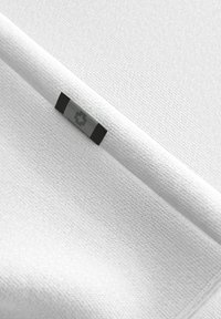 Weißer strukturierten Stoff mit flachem Gewebe, der ein kleines silbernes und schwarzes Etikett mit einem Logo aufweist. Das Material wirkt weich und leicht.