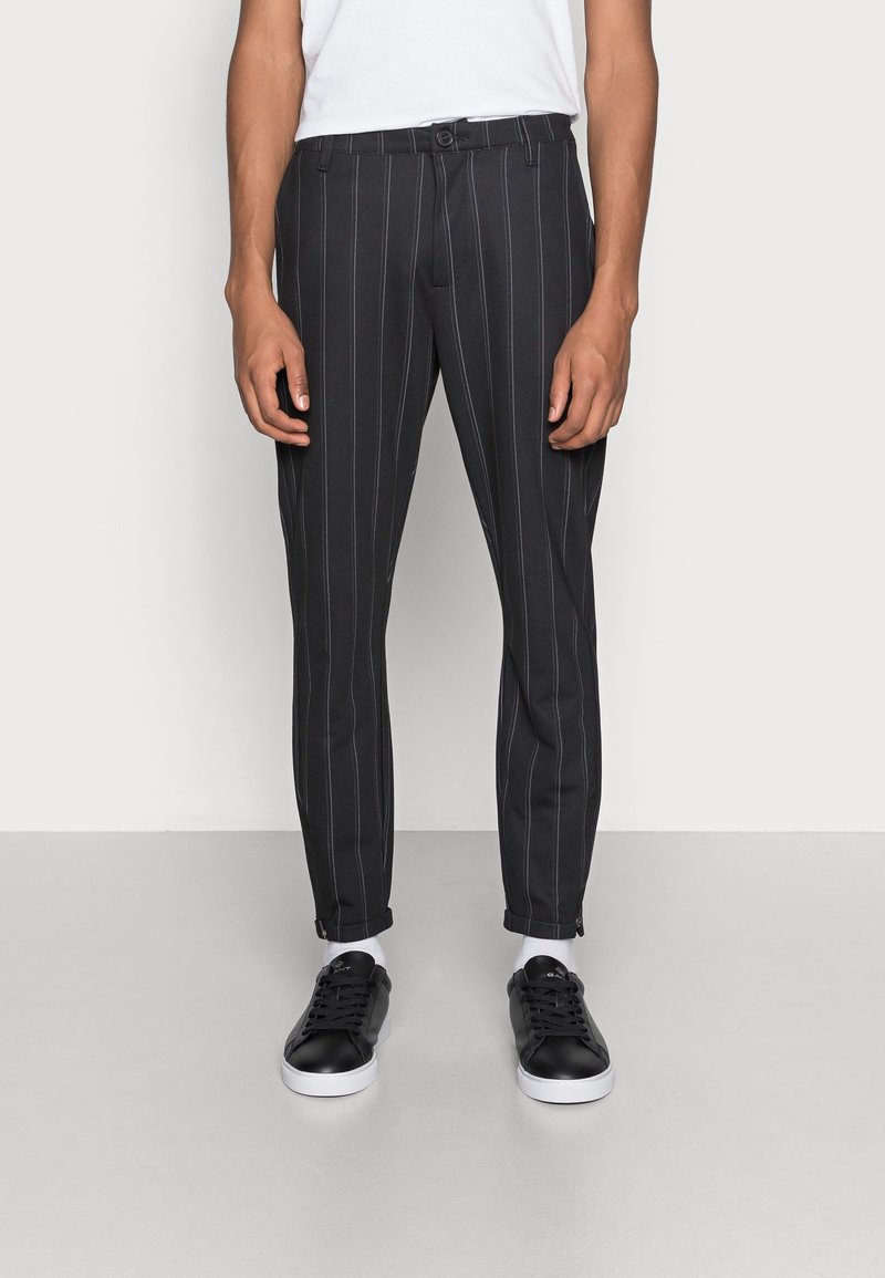 Mann trägt schwarze Slim-Fit-Hose mit weißen Nadelstreifen, kombiniert mit schwarzen Low-Top-Sneakern und einem weißen T-Shirt, steht vor einem einfarbigen Hintergrund.