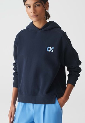 Felpa con cappuccio blu navy con logo frontale azzurro, caratterizzata da una vestibilità comoda e polsini a coste. Indossata con pantaloni azzurri.