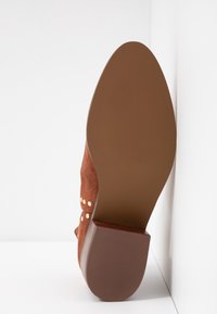 Bottine en suède marron avec un talon en caoutchouc marron épais. Présente des accents de clous décoratifs en or sur le côté et une semelle texturée.