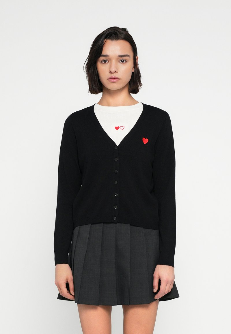 Μα ύρη ζακέτα με ντεκολτέ σε σχήμα V, κλείσιμο με κουμπιά και κεντημένη κόκκινη καρδιά στο στήθος, φορεμένη πάνω από λευκή μπλούζα και γκρι φούστα.