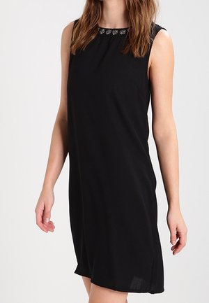 Ärmelloses, knielanges schwarzes Kleid mit rundem Ausschnitt und kleinen silbernen Blumenverzierungen, getragen von einer stehenden Person.