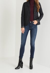 Czarna zamszowa marynarka z dwoma guzikami i kieszeniami, noszona na bordowym golfie. Ciemnoniebieskie rurki i błyszczące czarne botki na kostkę.