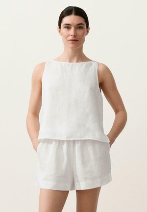 Žena s tmavými vlasmi v bielej bezrukávovej blúzke a zodpovedajúcich bielych šortkách stojaca so založenými rukami vo vreckách pred jednoduchým pozadím.