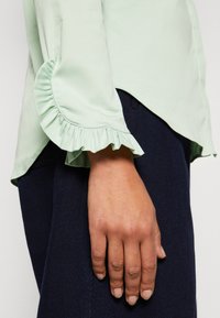 Main reposant sur le côté portant un chemisier vert clair à manches longues avec des poignets à volants et un pantalon bleu foncé.