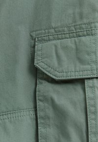 Tissu vert olive avec une texture lisse, présentant une poche de poitrine proéminente et des détails de couture visibles le long des coutures.