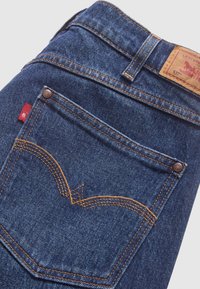 Vaqueros de mezclilla azul oscuro con costuras naranjas. Presentan una etiqueta de cuero con logotipo y un pequeño parche rojo en el bolsillo trasero con un diseño distintivo.