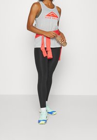 Graues Netz-Trägershirt mit orangem "Nike Trail"-Logo, schwarze Leggings und blau-grüne Laufschuhe. Rote Jacke um die Taille gebunden. Hält eine Wasserflasche in der Hand.