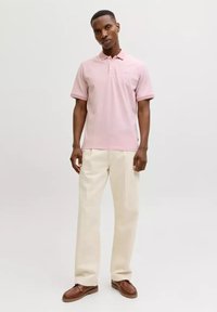 Mann trägt leichtes rosa Polohemd mit kurzen Ärmeln, cremefarbene Hose und braune Lederschuhe, steht vor einem schlichten weißen Hintergrund.