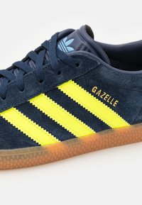 Námořnické modré semišové tenisky s třemi neonově žlutými pruhy, zlatým nápisem "Gazelle" a světle modrým logem. Obsahují gumovou podrážku.