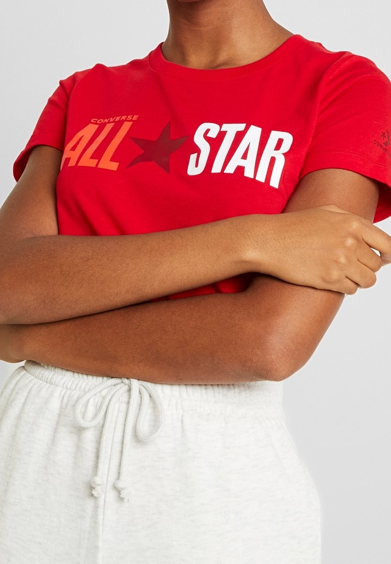 Personne portant un t-shirt rouge Converse All Star avec texte blanc et pantalon blanc à cordon, les bras croisés sur la poitrine.