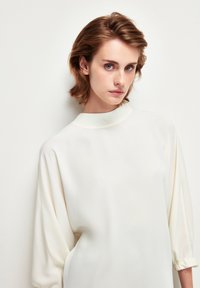 Blusa bianca in seta con un collo alto, maniche morbide raccolte ai polsi e una texture liscia. Modella mostrata su uno sfondo chiaro.