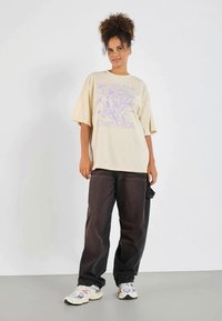 T-shirt beige surdimensionné avec un graphique floral violet et le texte "Own Pace," associé à un pantalon noir ample et des baskets multicolores.