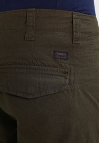 Olivgröna byxor med texturerad yta, med en bakficka och en märkeslapp. Stickade detaljer syns vid midjan.