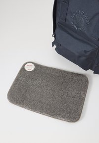 Grå sittdyna med en texturerad yta, rektangulär form, placerad på en vit bakgrund bredvid en mörk ryggsäck. Har en cirkulär etikett.